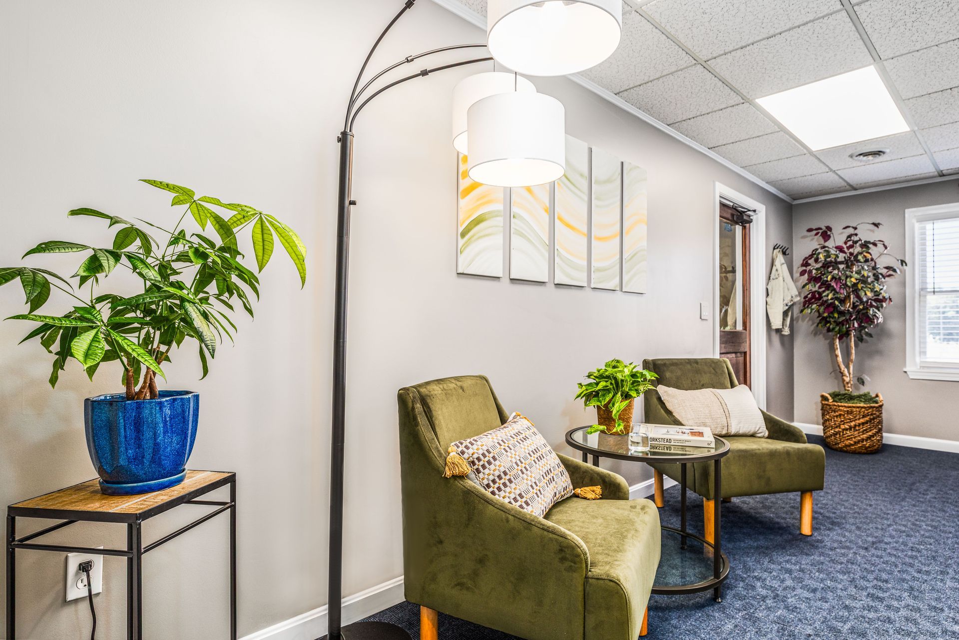 A waiting room with two olive armchairs, a floor lamp, potted plants, wall art, and blue carpeted flooring.