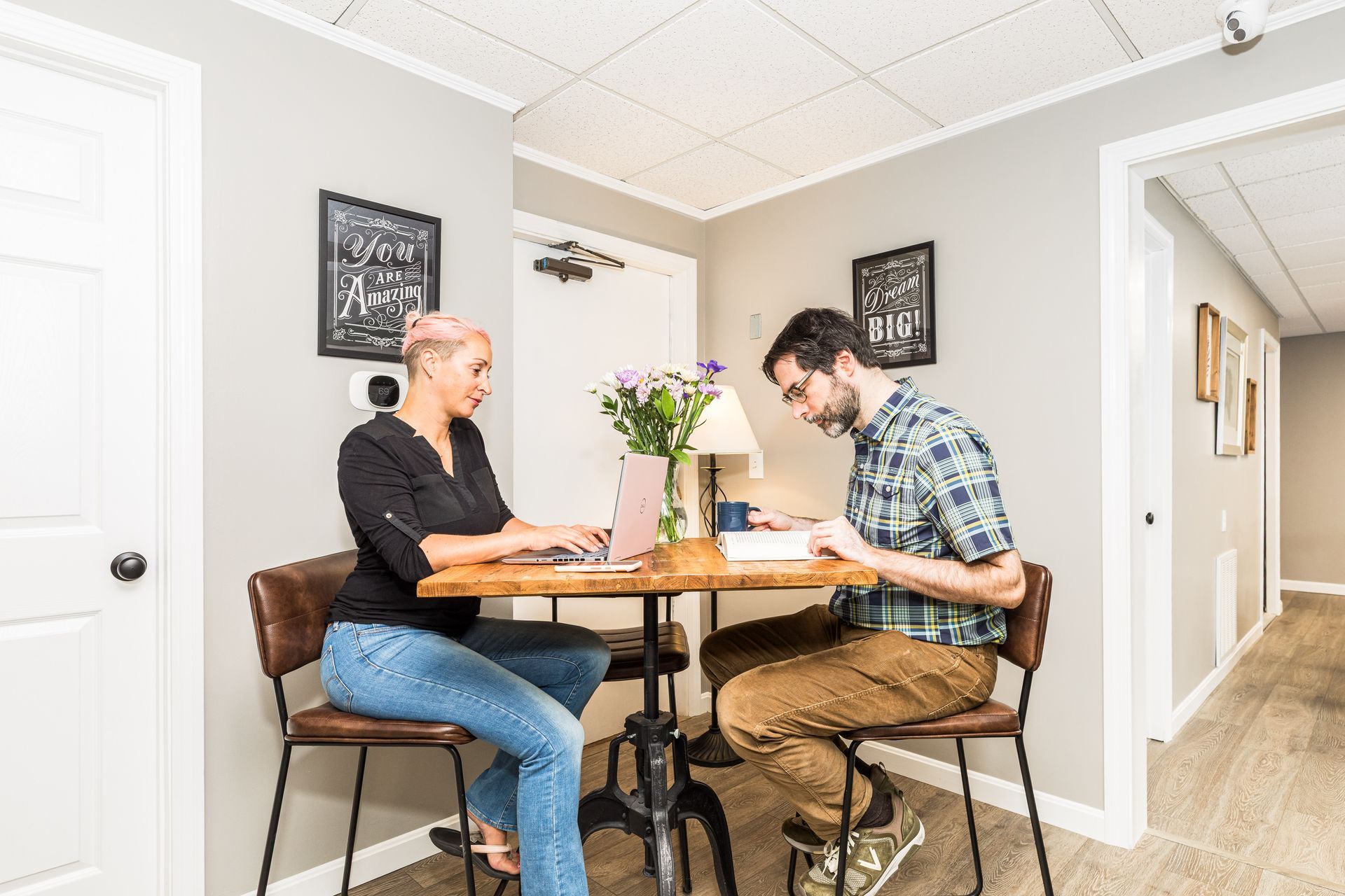Two people sit at a wooden table in an office setting, working on a laptop and reading a document.