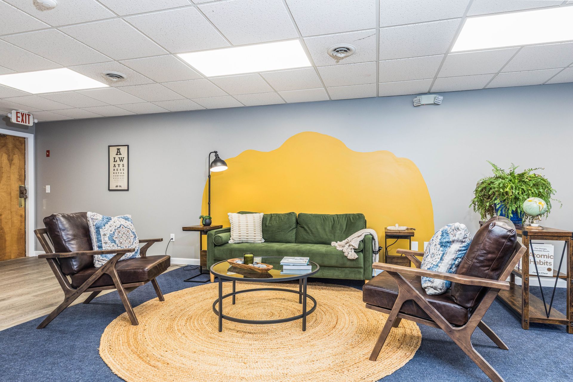Cozy office lounge with a green couch, two leather chairs, a central rug, and a yellow painted cloud accent wall.