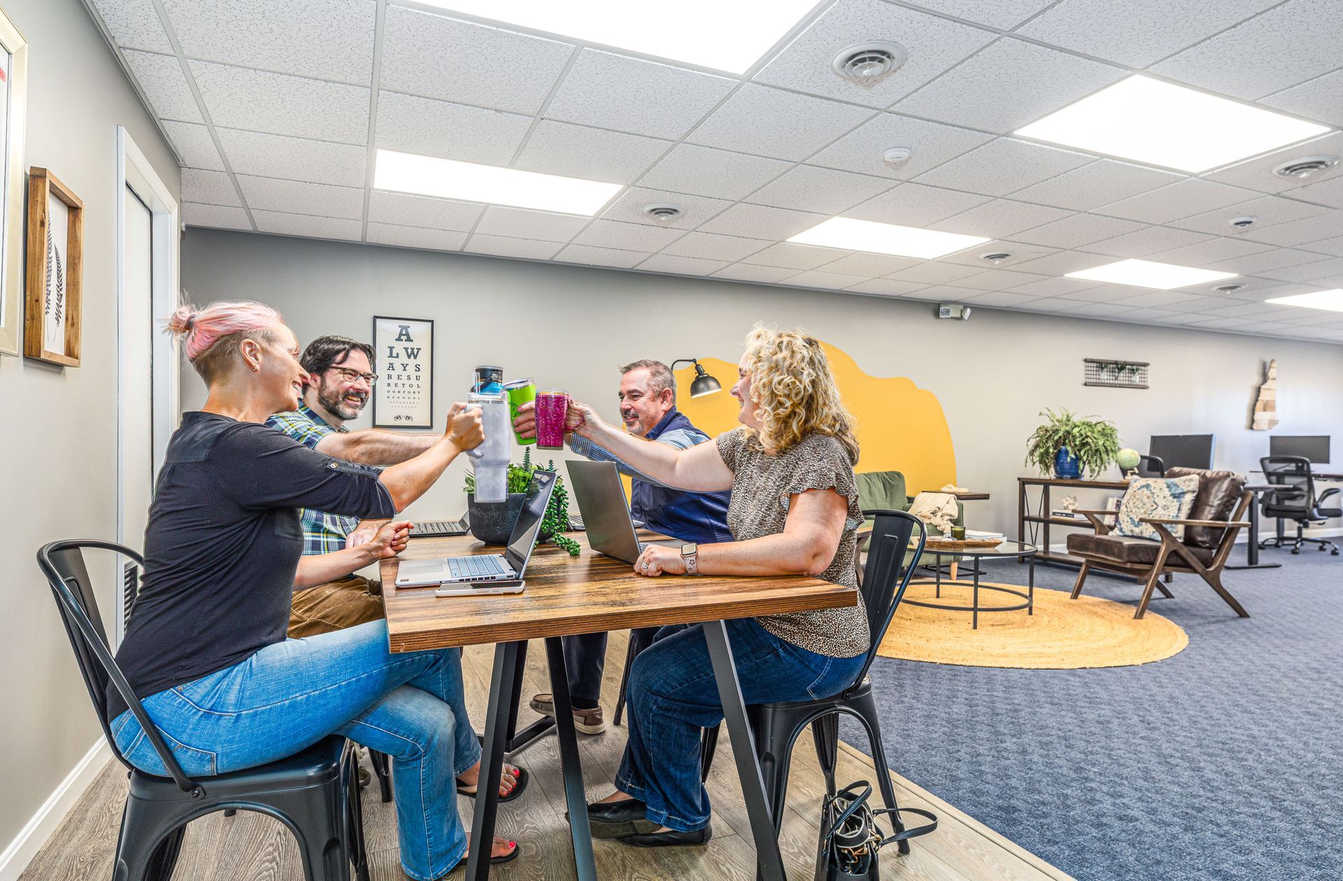 Four people toast with beverages around a wooden table in an office setting.