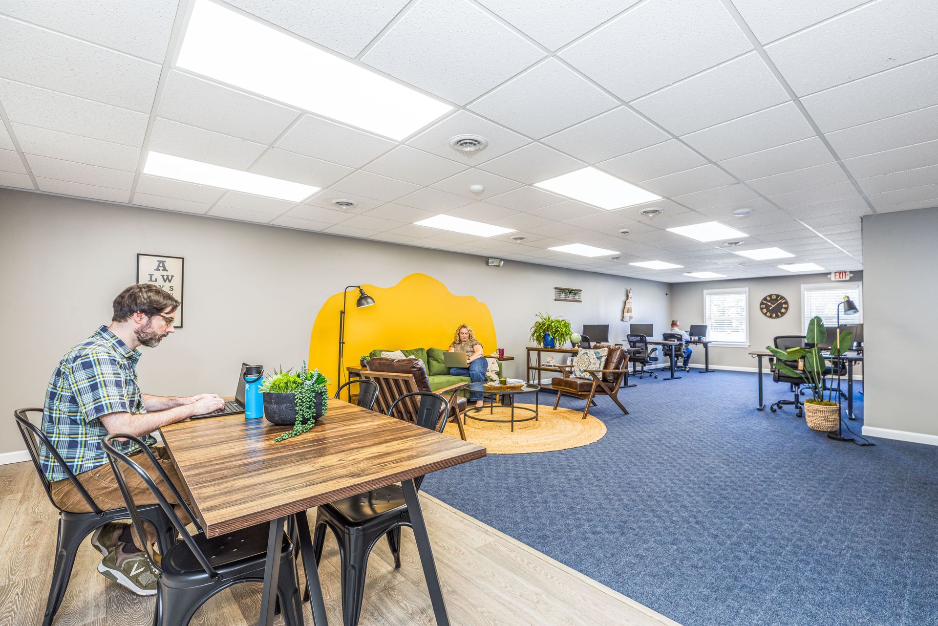 A person works on a laptop at a wooden table in a brightly lit, open-plan office with a yellow accent wall and blue carpet.