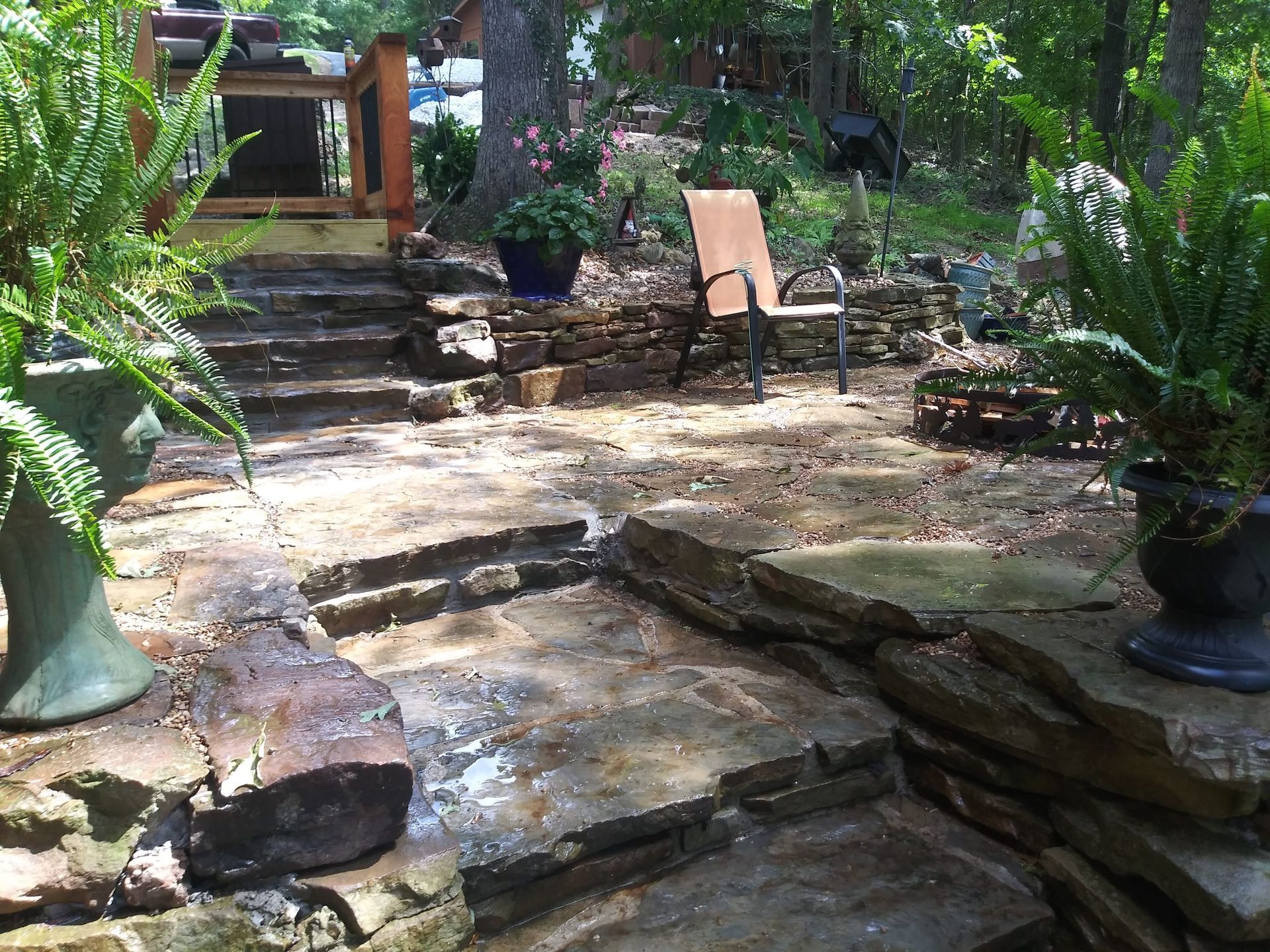 Stone patio with ferns in urns, stairs, and a chair in a wooded area.