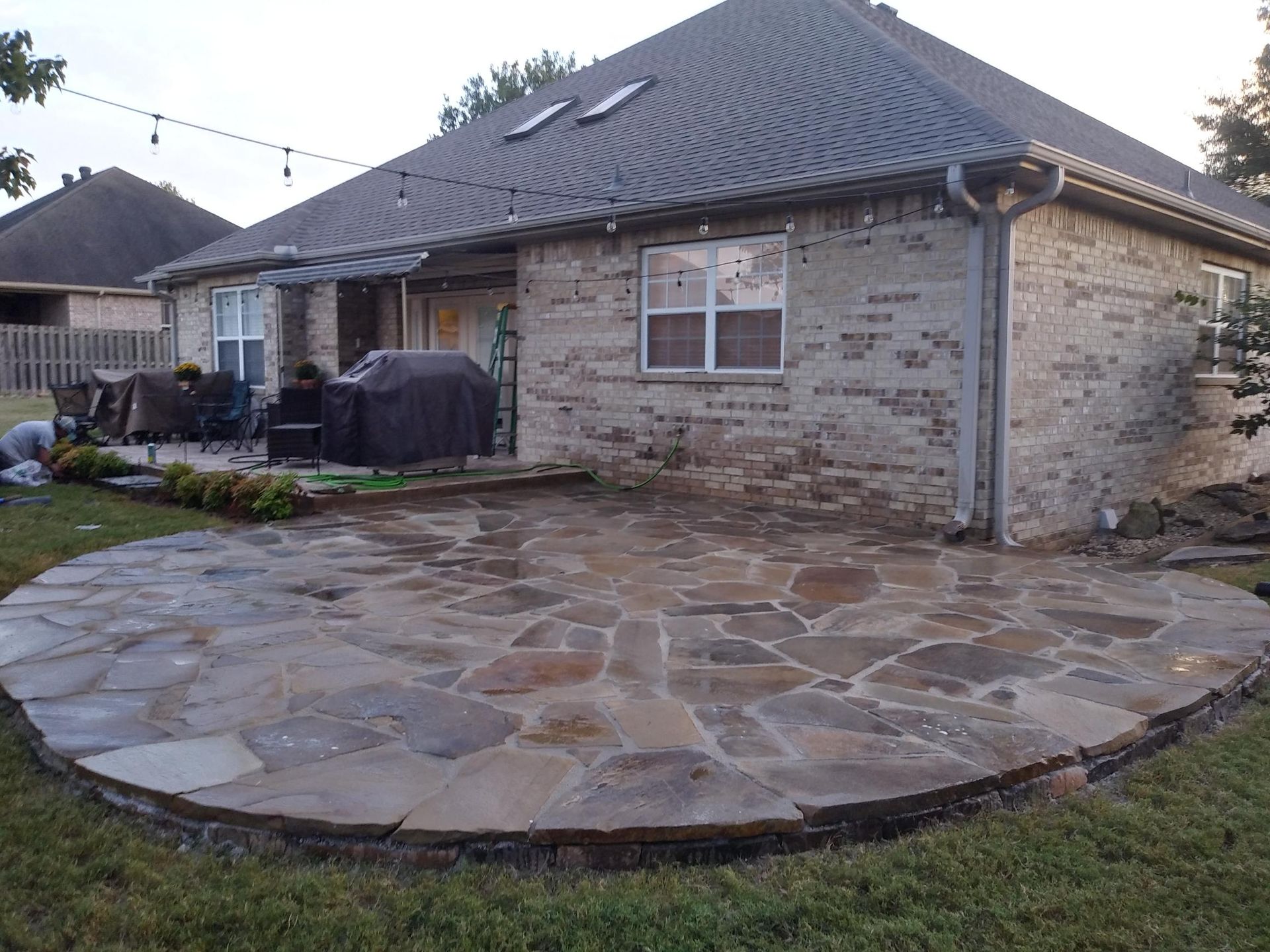 Stone patio next to a brick house, surrounded by grass. String lights above the patio.
