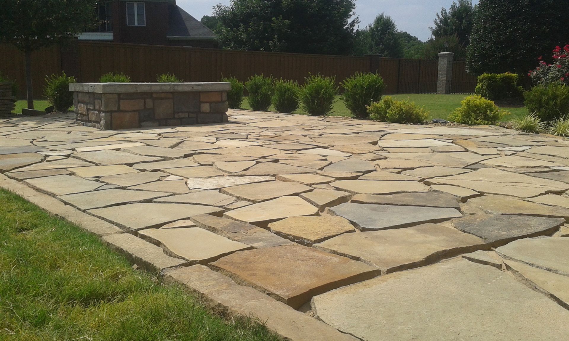 Stone patio with fire pit surrounded by green grass and shrubs.