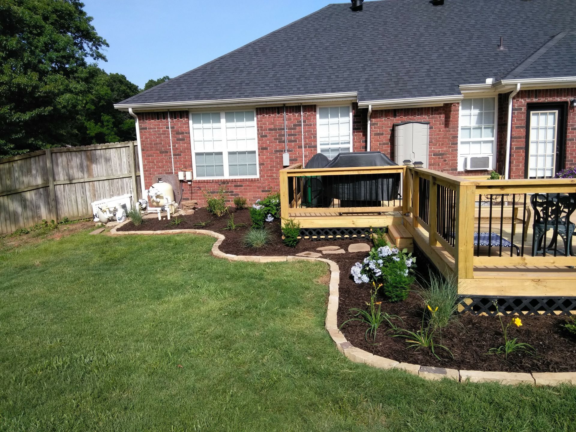 Backyard with a brick house, wooden deck, and garden beds filled with flowers.