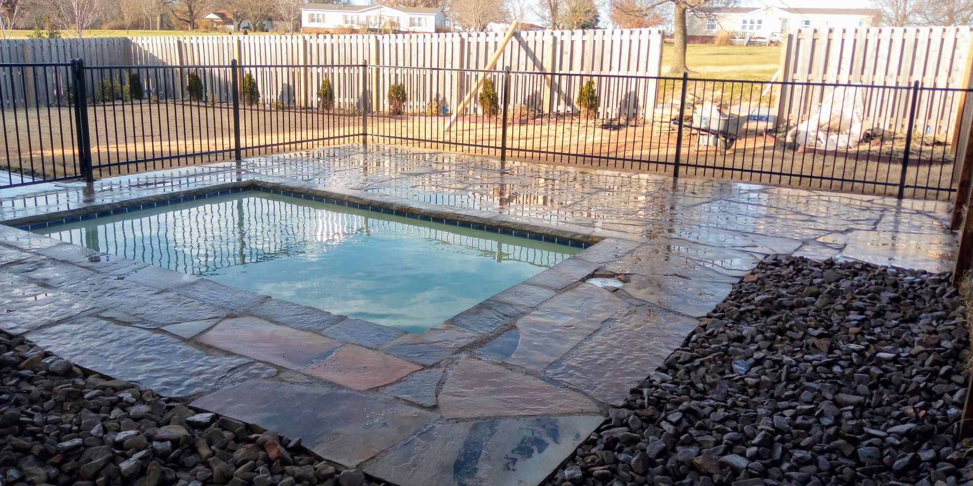 Small pool surrounded by stone patio and black fence, with gravel ground cover.