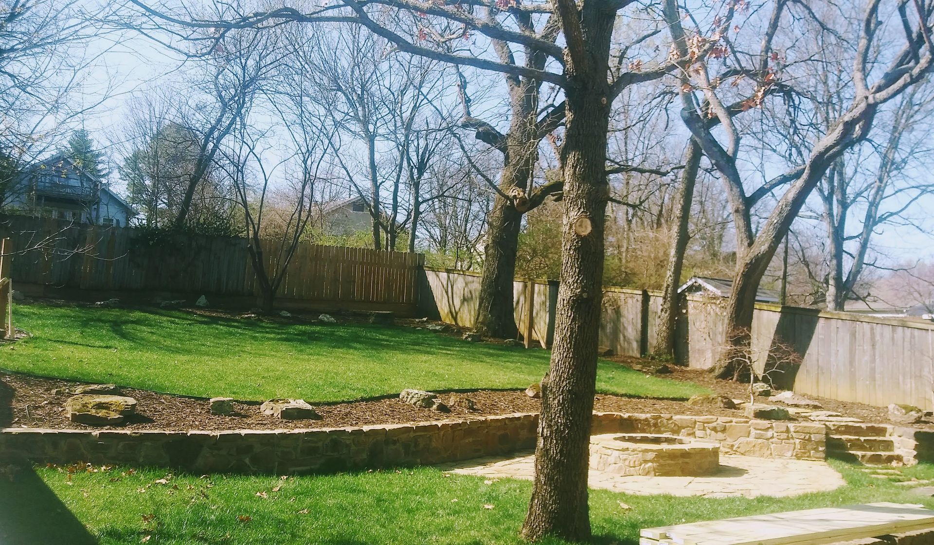Grassy backyard with a retaining wall, trees, and a wooden fence on a sunny day.