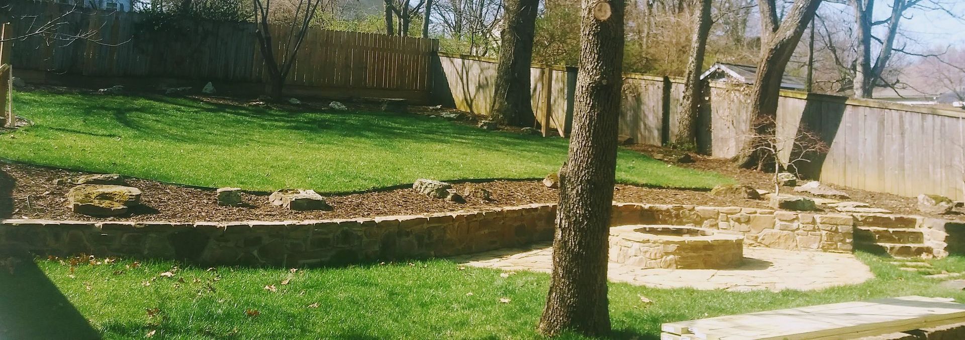 Backyard with grass, trees, retaining walls, and a fence on a sunny day.