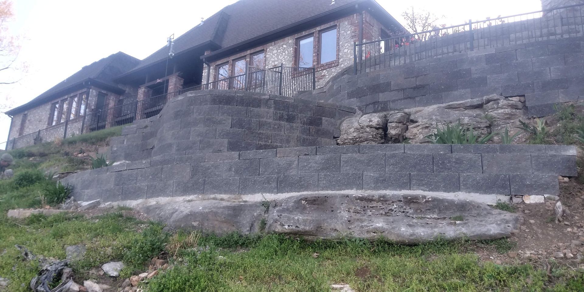 A stone house on a hill, built with retaining walls, and staircases. Green grass and overcast sky.