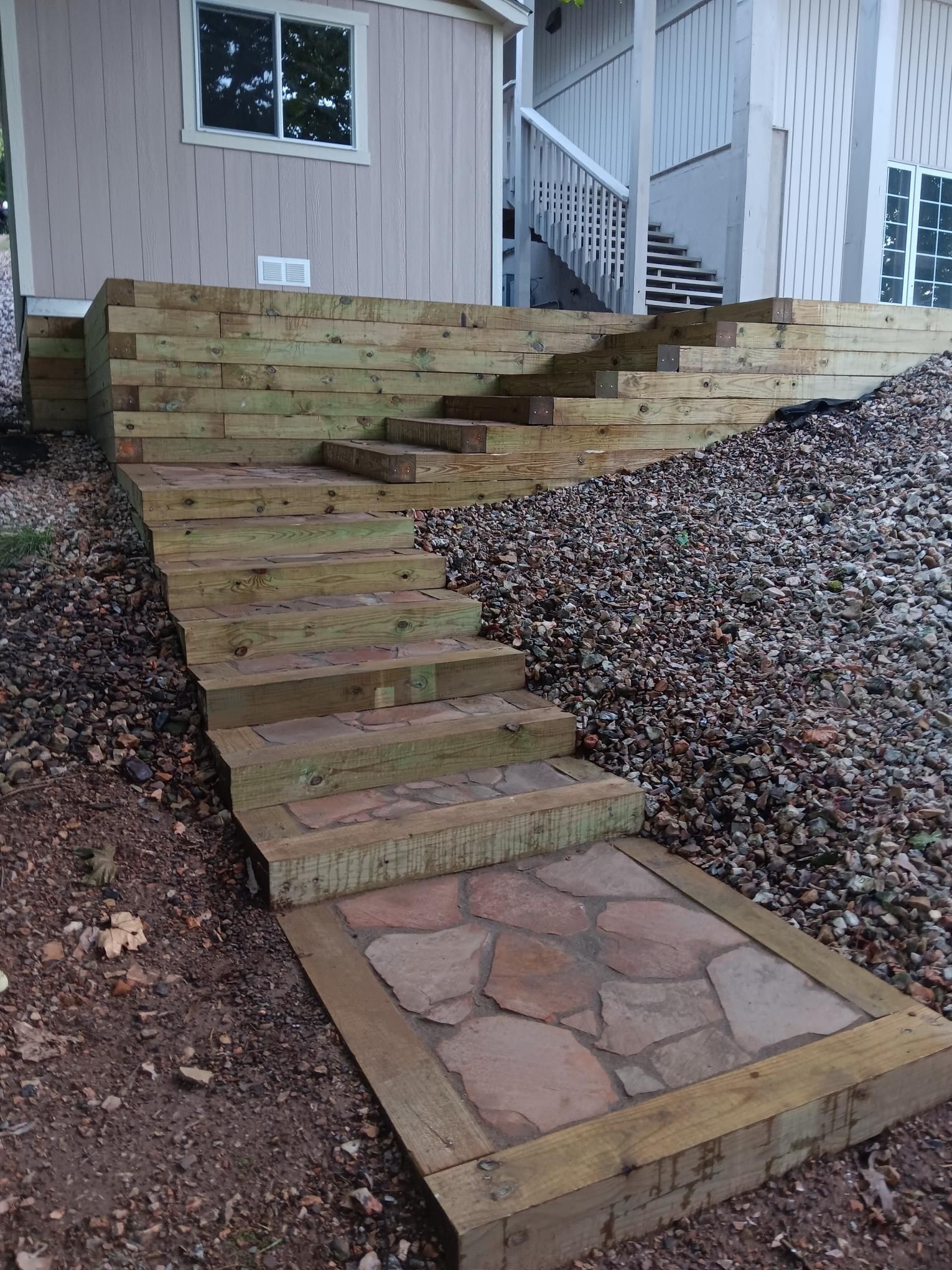 Wooden steps leading up a hillside, stone patio at the base, gravel and dirt surround.