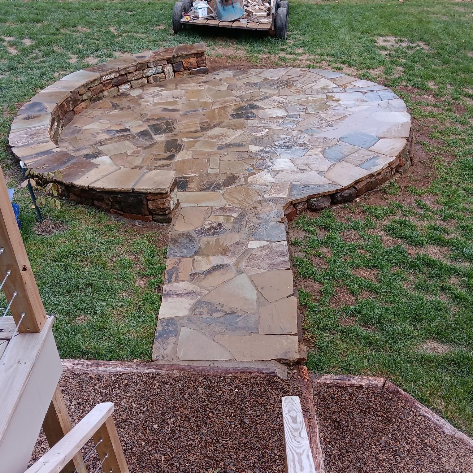 Stone patio with curved seating wall, connected by a stone path to a lower level, on a green lawn.