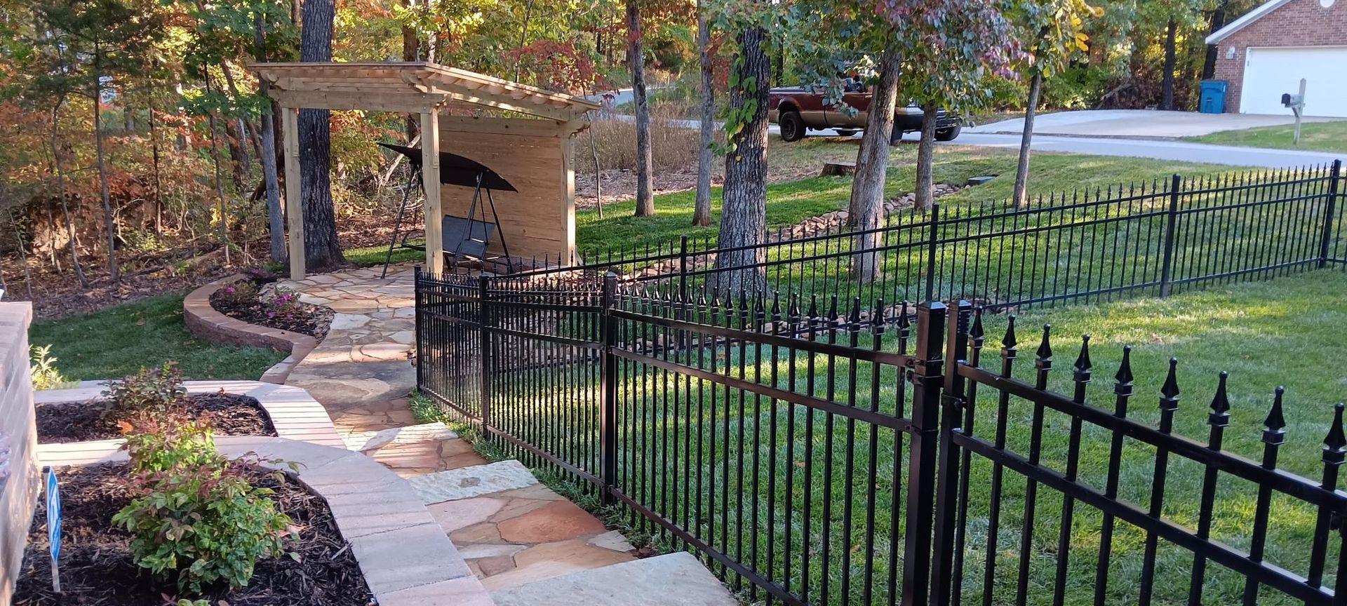 Black metal fence lines a stone walkway leading to a structure in a yard. Trees and grass surround it.