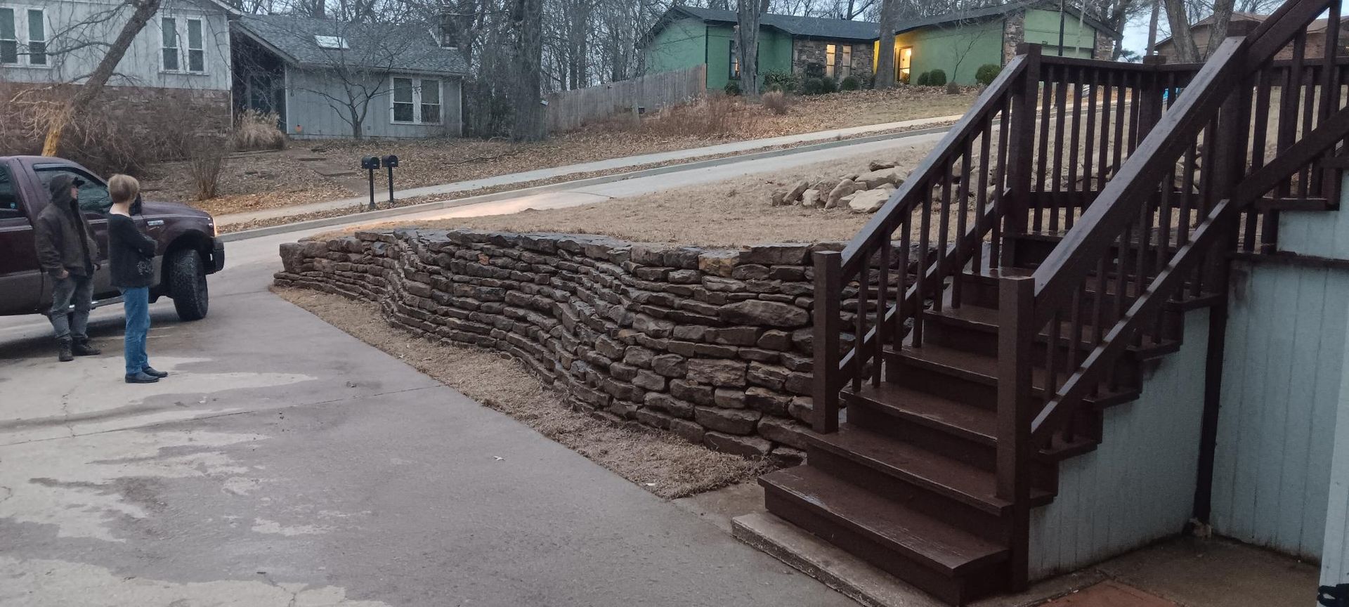 People standing near a truck in a driveway next to a stone wall and stairs leading up to a house.