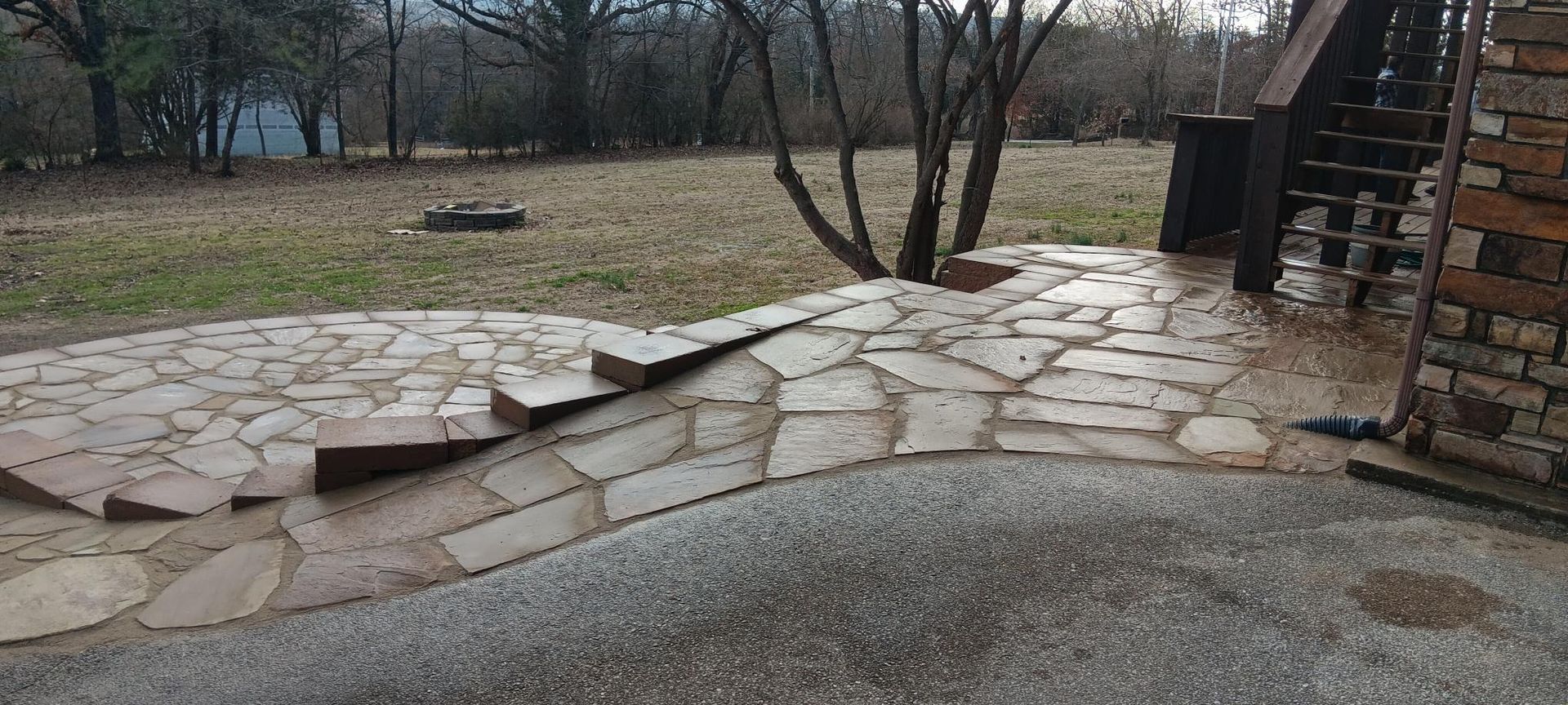 A stone patio with a ramp leading up to a yard, brick building to the right. Bare trees in the background.
