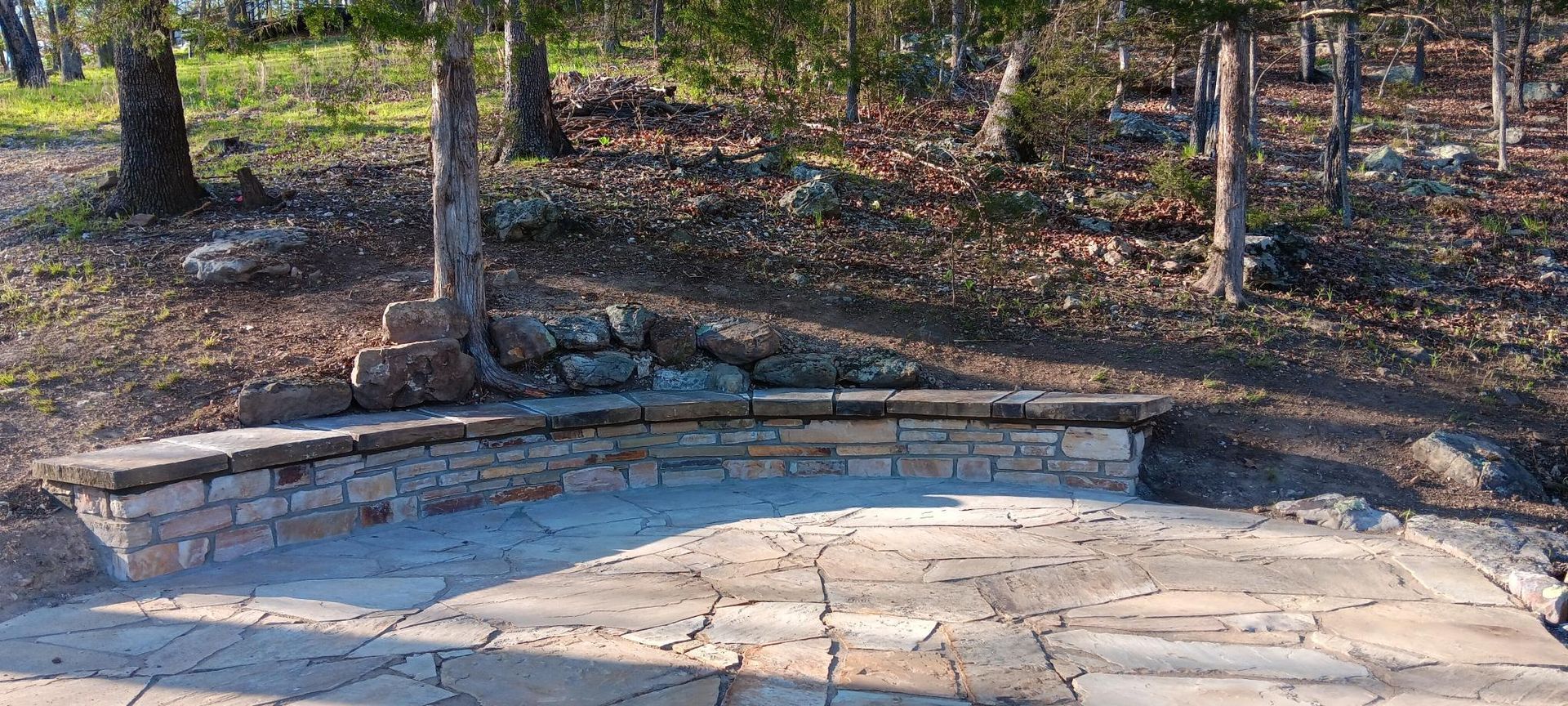 Stone patio with a stone wall in a wooded area. Trees and foliage in the background.