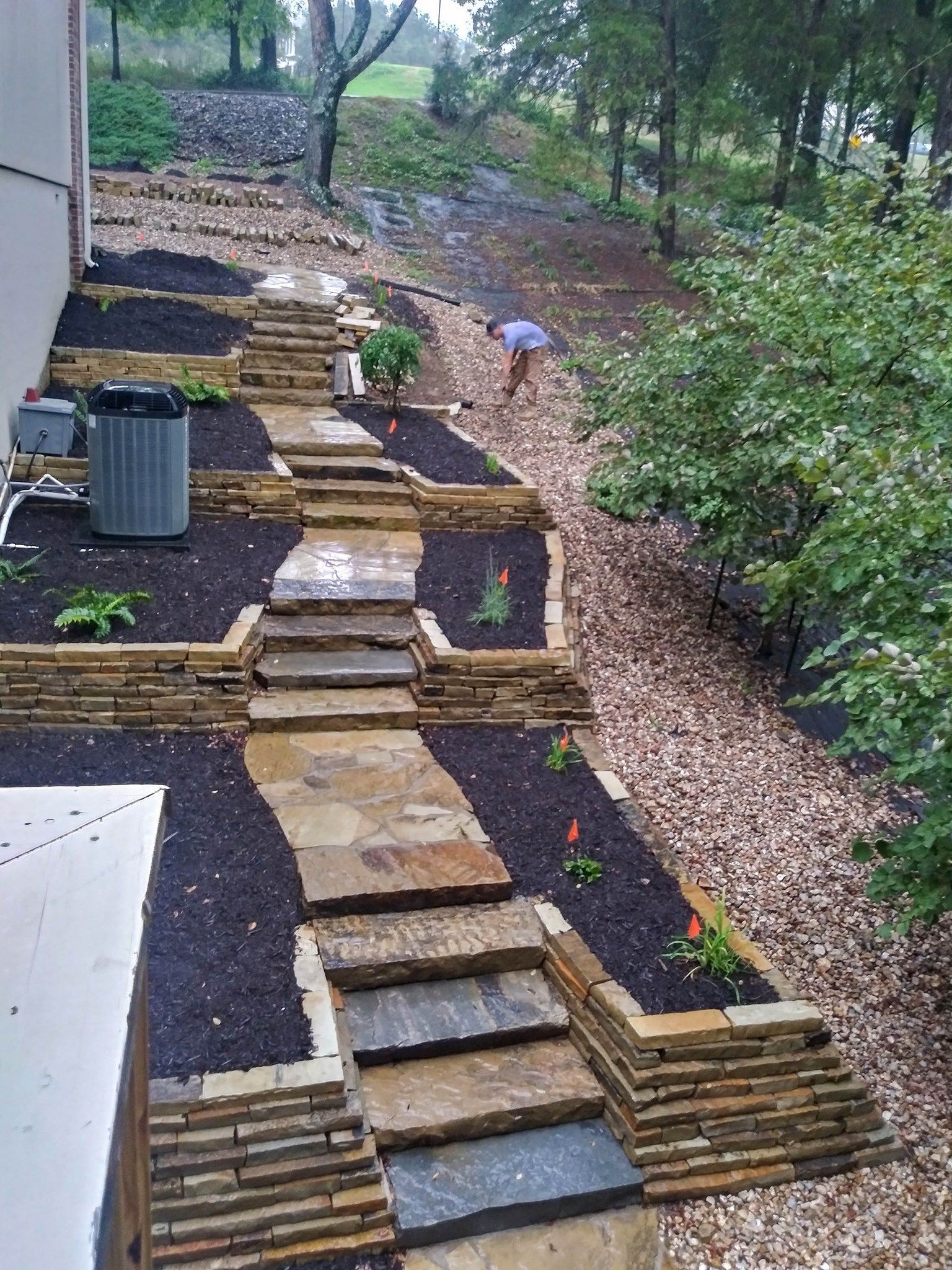 Stone steps and retaining walls ascend a hillside, landscaped with dark mulch and plants. A person works on the upper path.
