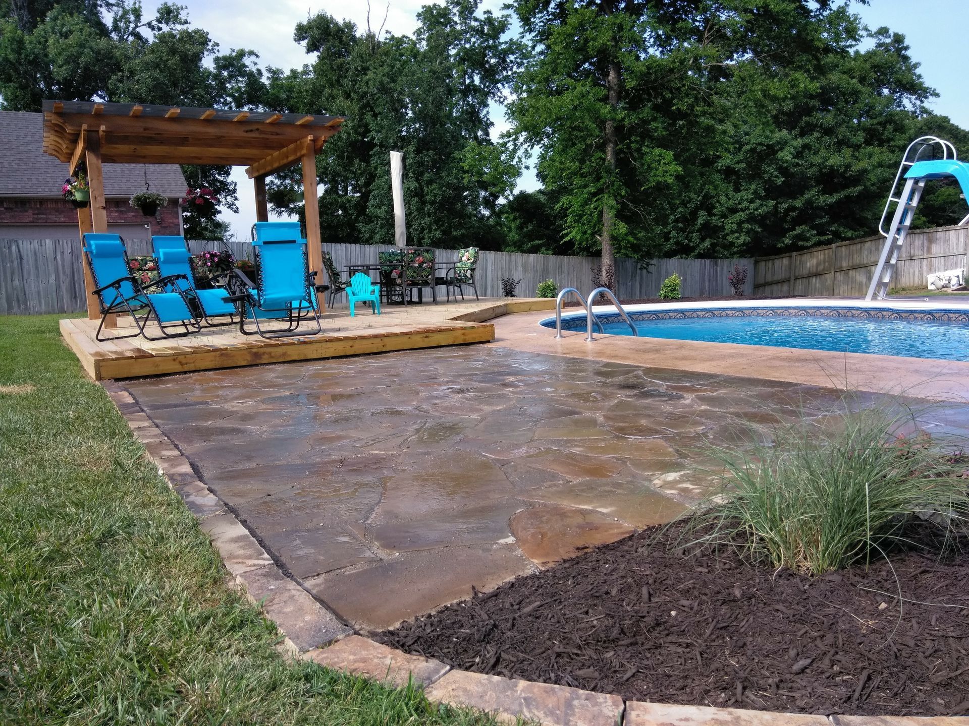 Backyard pool area with wooden pergola and blue lounge chairs on a wooden deck.