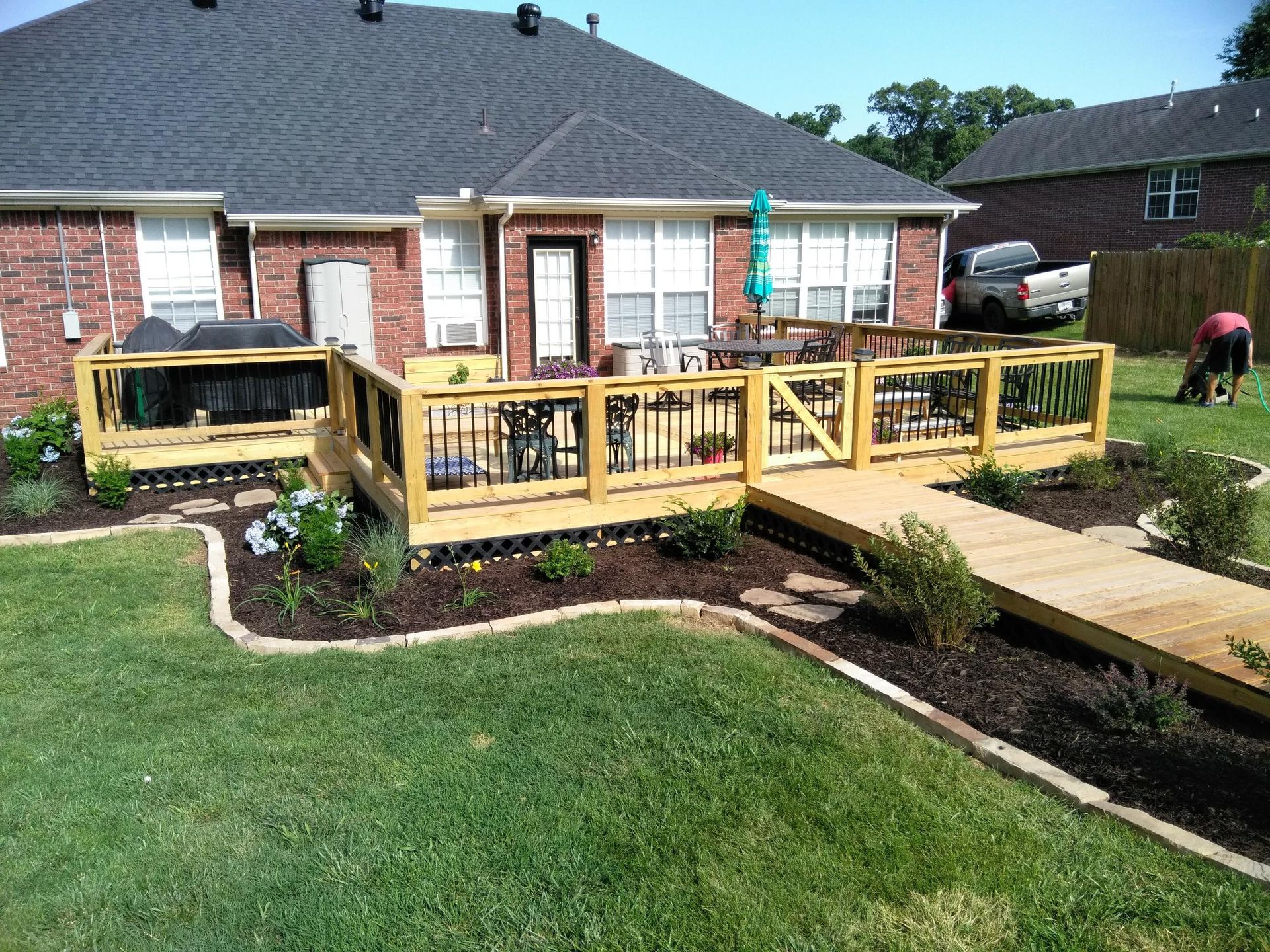 Backyard deck with wooden ramp, black railings, brick house, green grass, and garden beds.