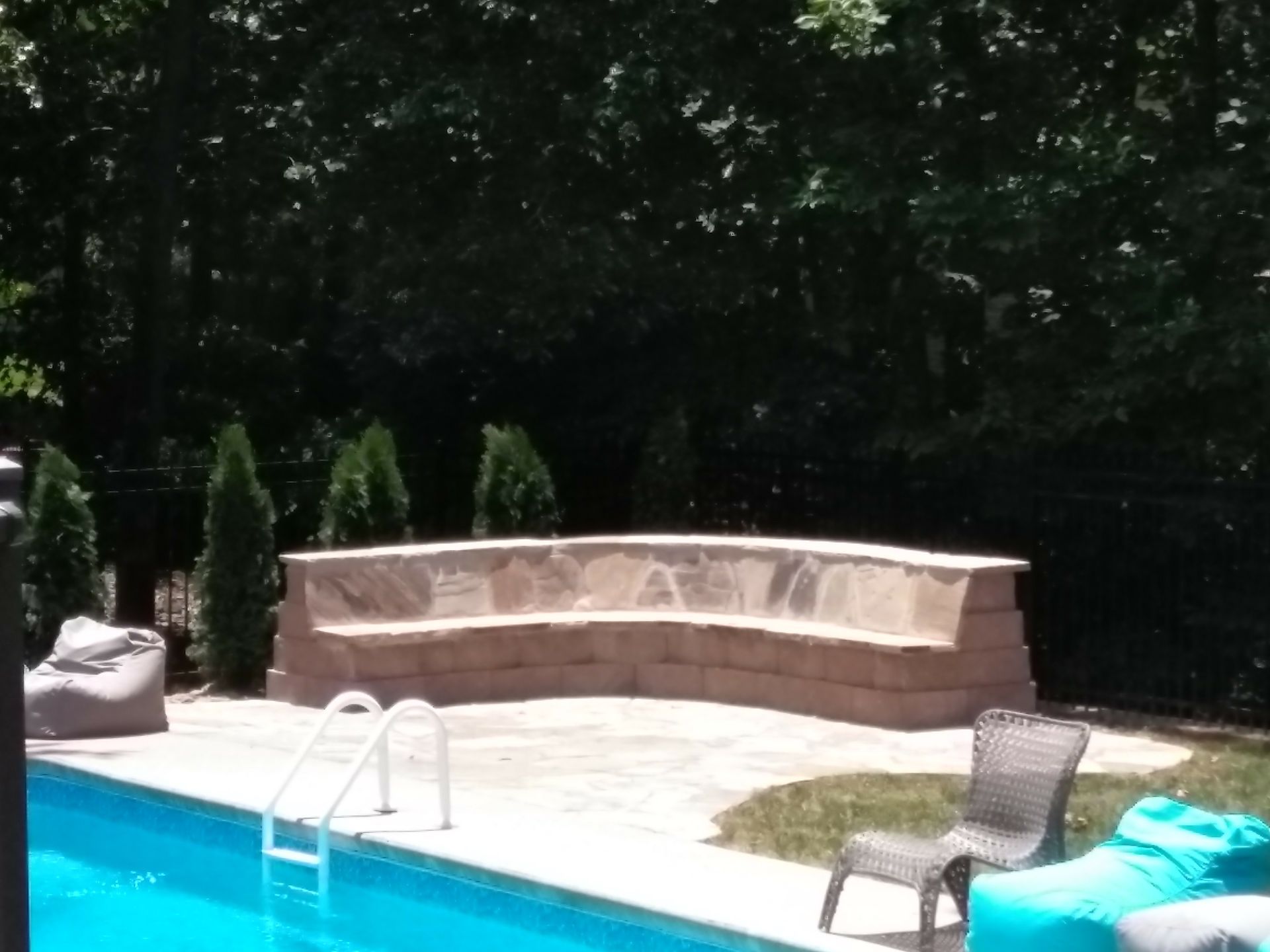 Stone bench seating by a pool, with trees in the background. Bright blue pool water.