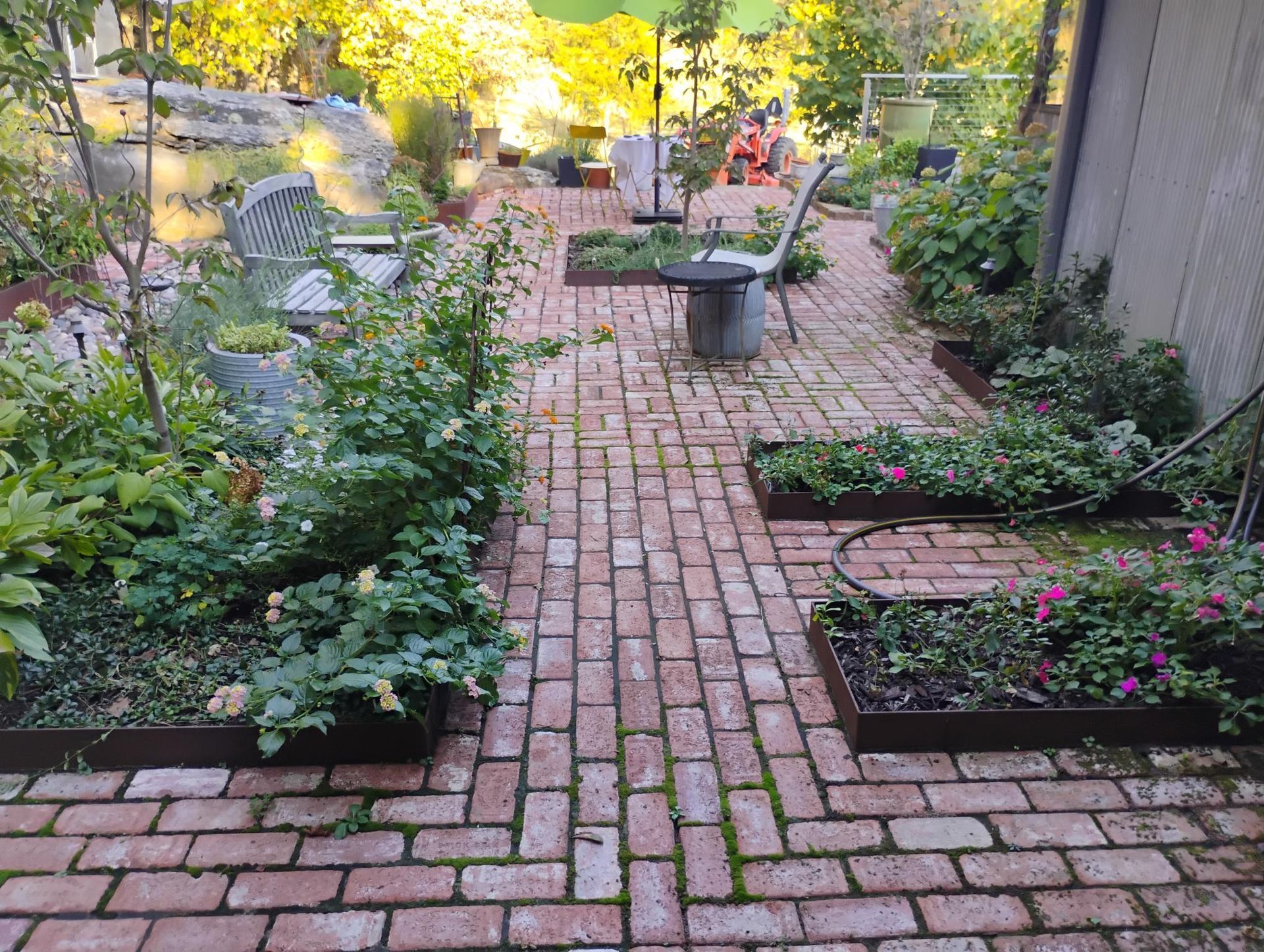 Brick patio with raised garden beds and lush plants. A small table and chairs are in the distance.