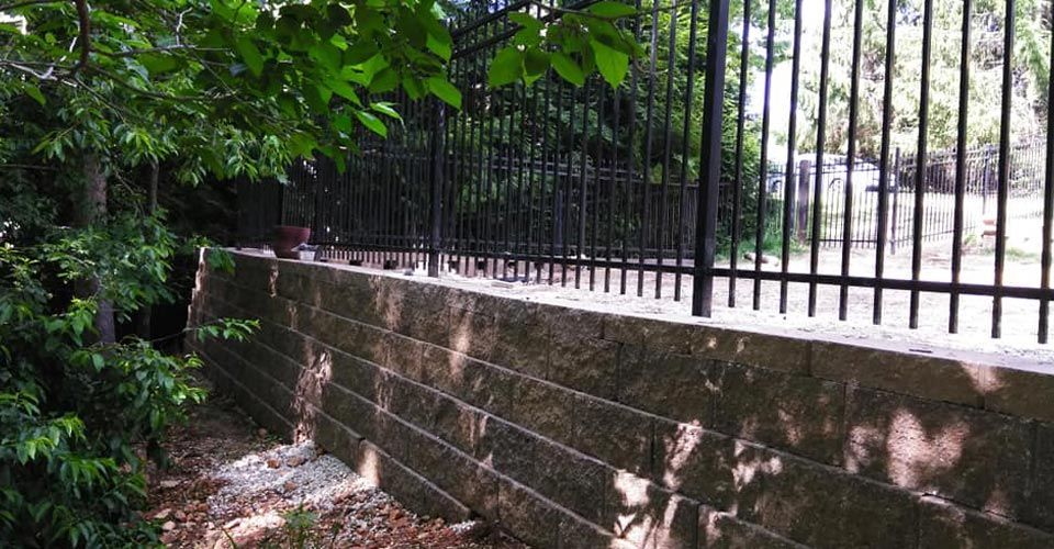 Black metal fence atop a retaining wall, with trees to the left.