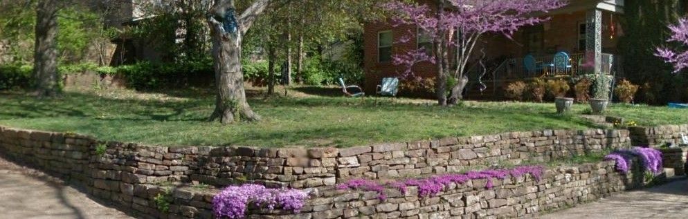 Stone wall with pink flowers and a green lawn, trees, and building in the background.