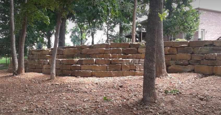 Stone retaining wall in a yard, with trees in front and a building in the background.