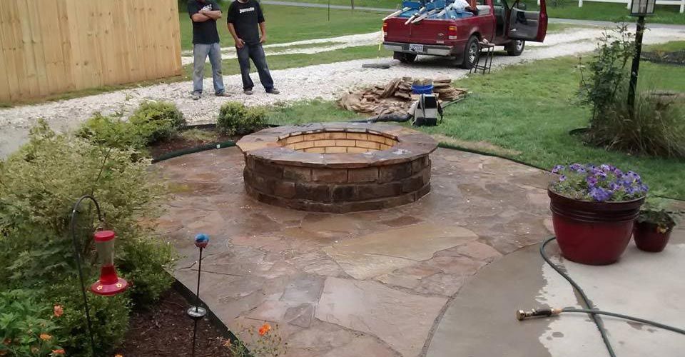 Fire pit on stone patio with two men, truck, and garden.