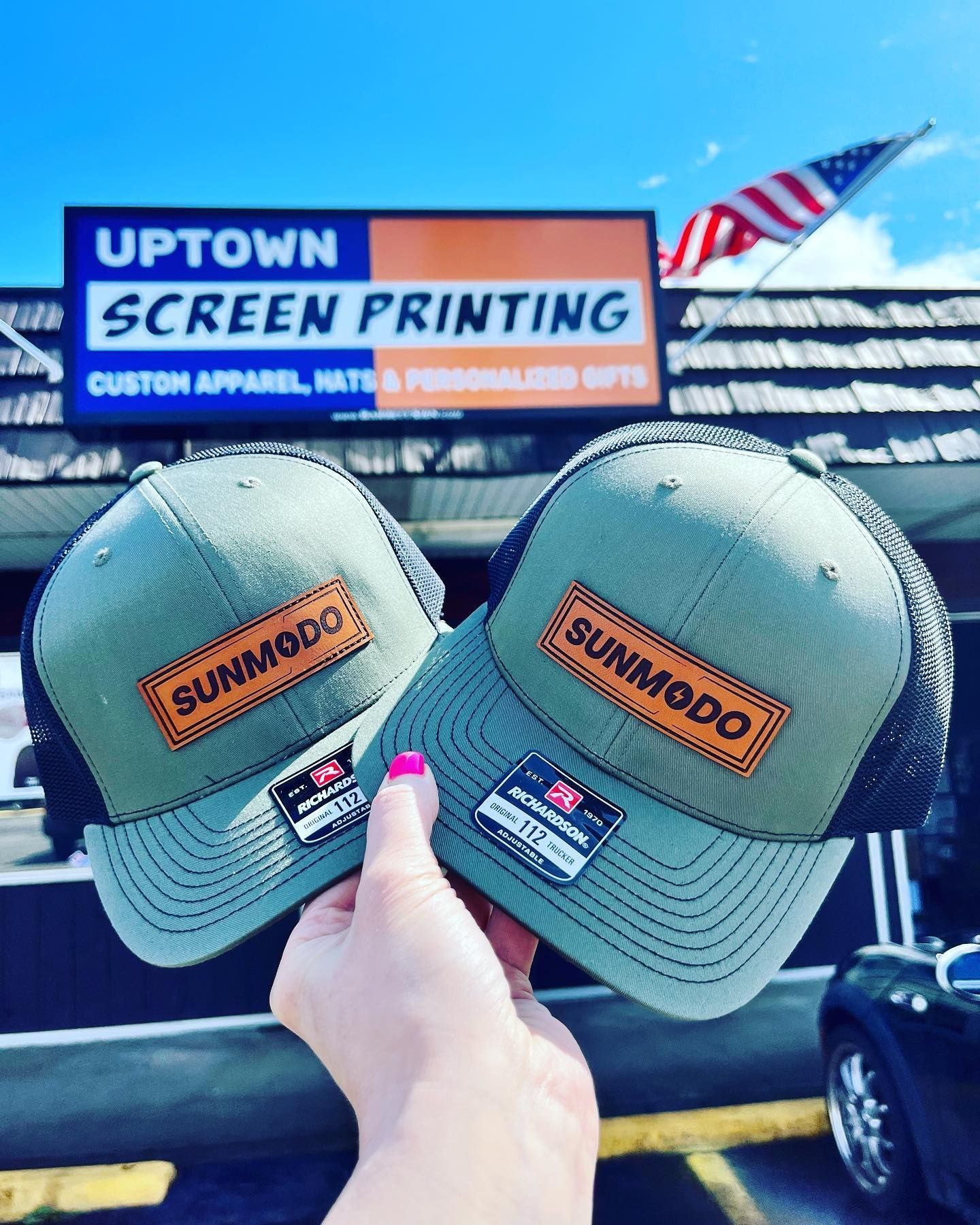 A person is holding two hats in front of a store called uptown screen printing