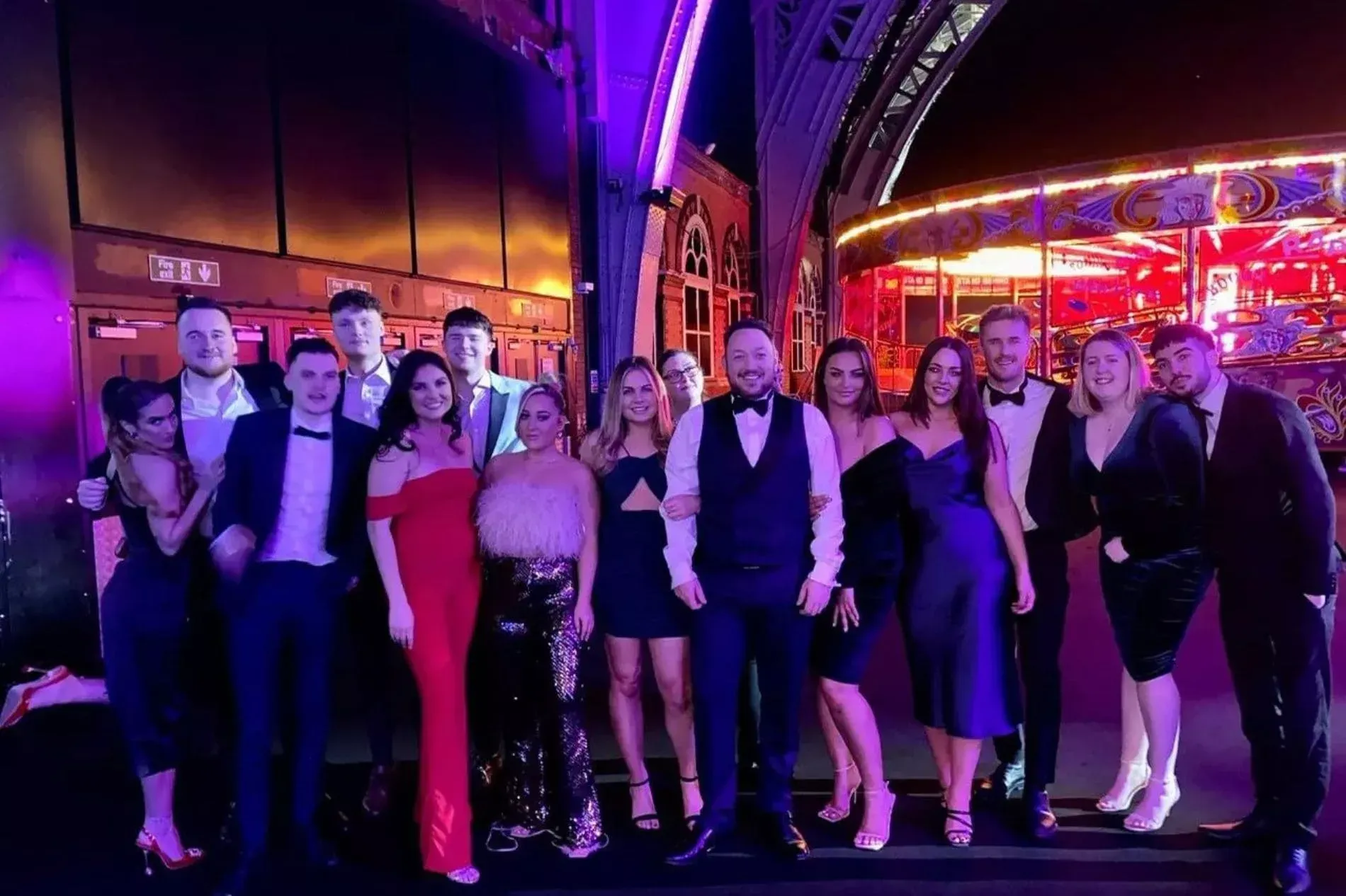 Group of formally dressed people smiling for the camera, lit with purple light, with a carousel in the background.