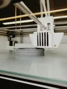 A 3D printer is building a gray, circular object on a white platform. The printer head is white.