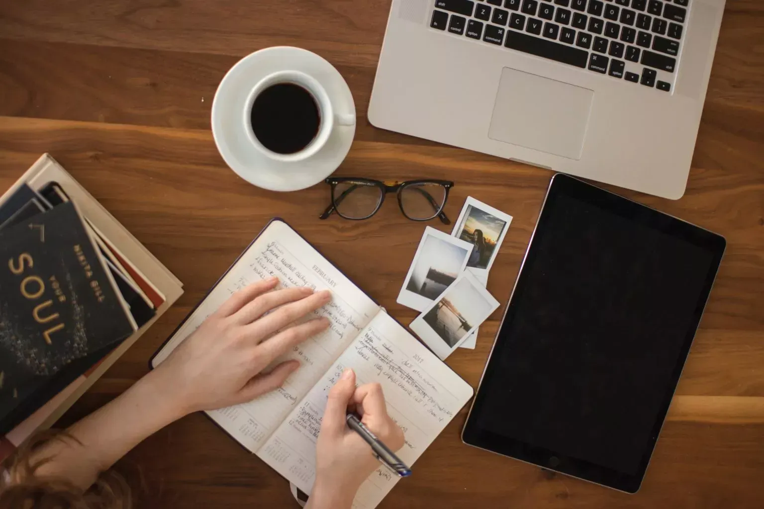 Person writing in a notebook on a wooden desk with a laptop, coffee, books, glasses, and photos.
