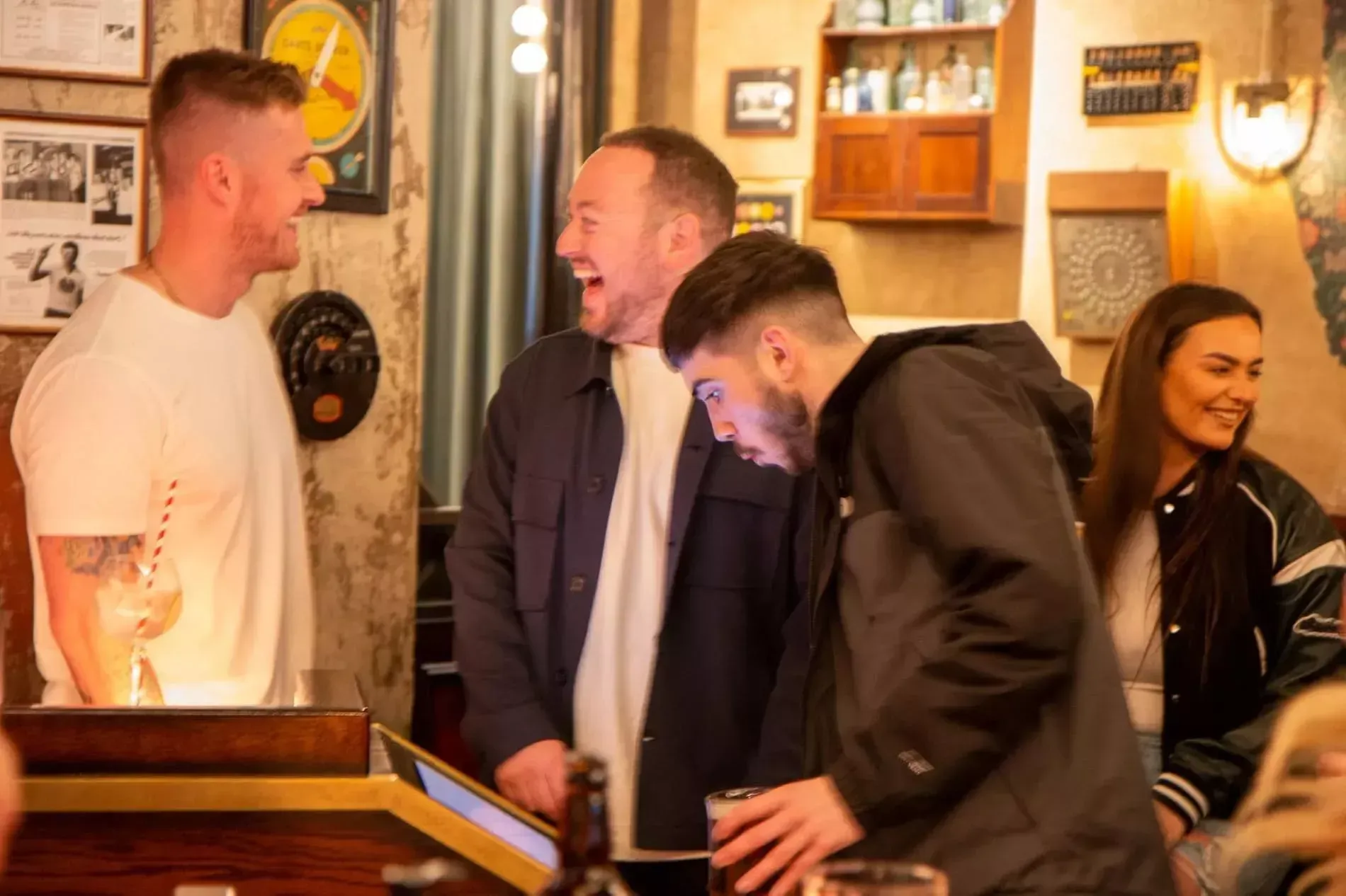 Group of friends laughing in a pub, near a bar. One person is looking at a phone. Warm lighting.