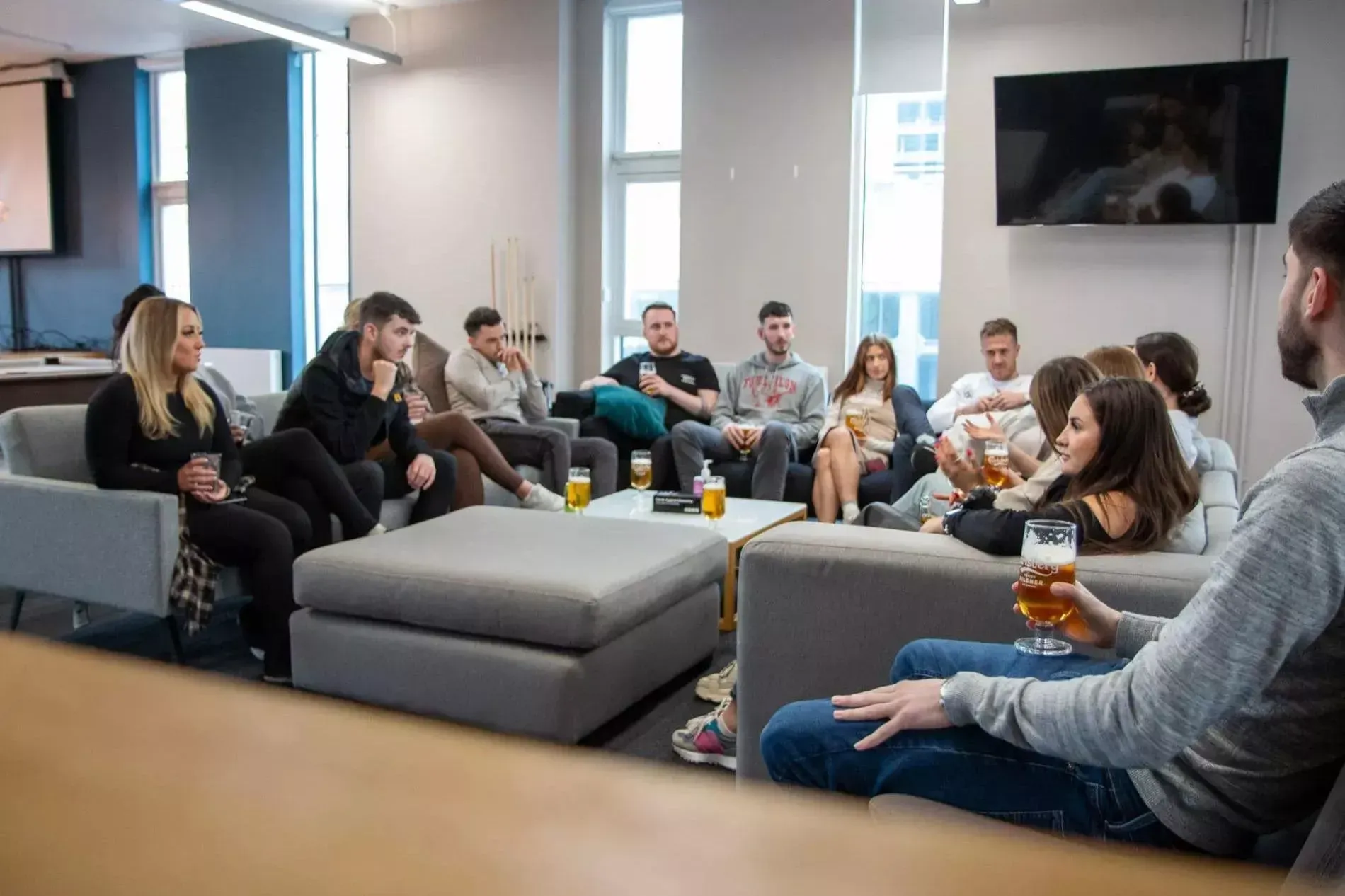 A group of young adults seated on sofas, socializing, drinking, in an office-like setting.