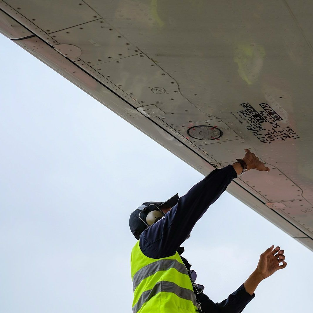 Aircraft mechanic inspecting an airplane wing, wearing a yellow safety vest and headset.