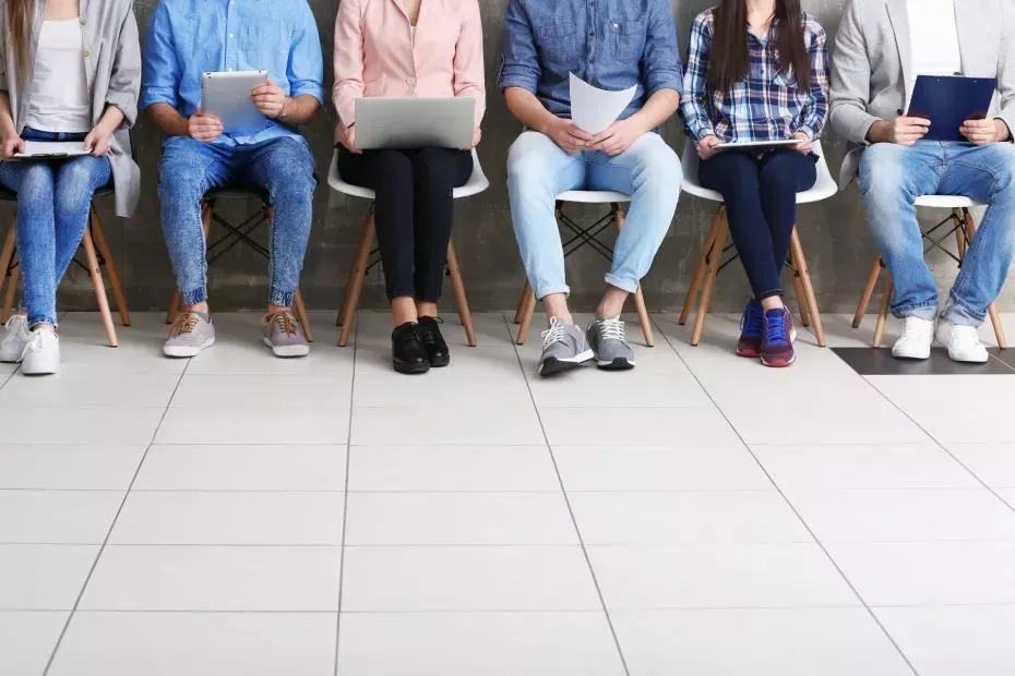 People seated in a row, holding devices, waiting. Jeans, shirts, shoes, against a gray wall.
