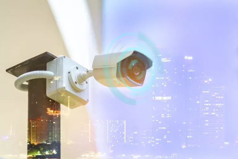 Security camera mounted outdoors with city background, blue light effect.