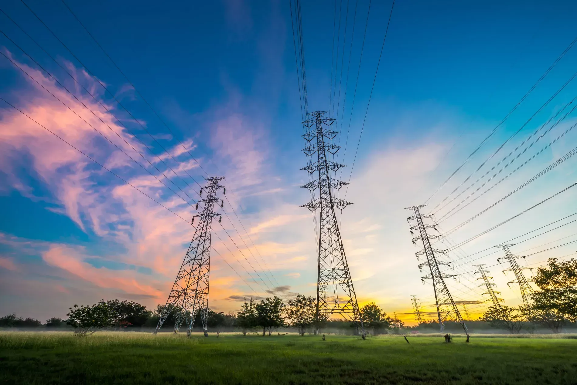 Power lines on tall towers crossing a green field under a colorful sunset.