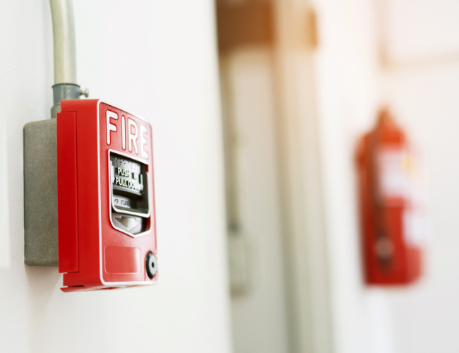 Red fire alarm on a white wall, fire extinguisher blurred in the background.