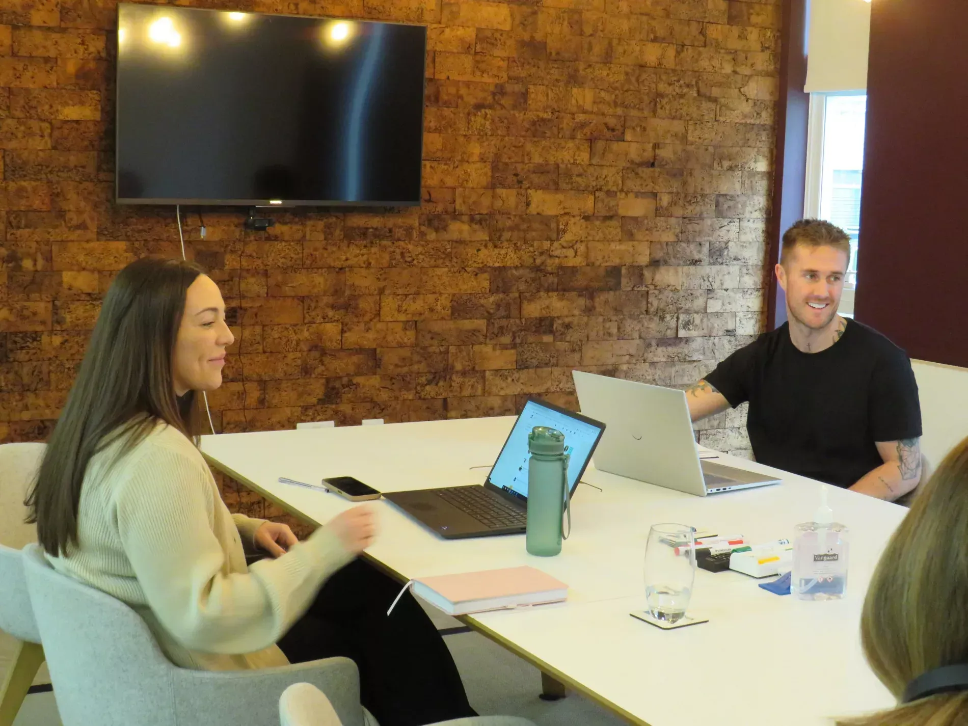 Two people, a man and a woman, at a table with laptops and a TV on the wall. Both are smiling.