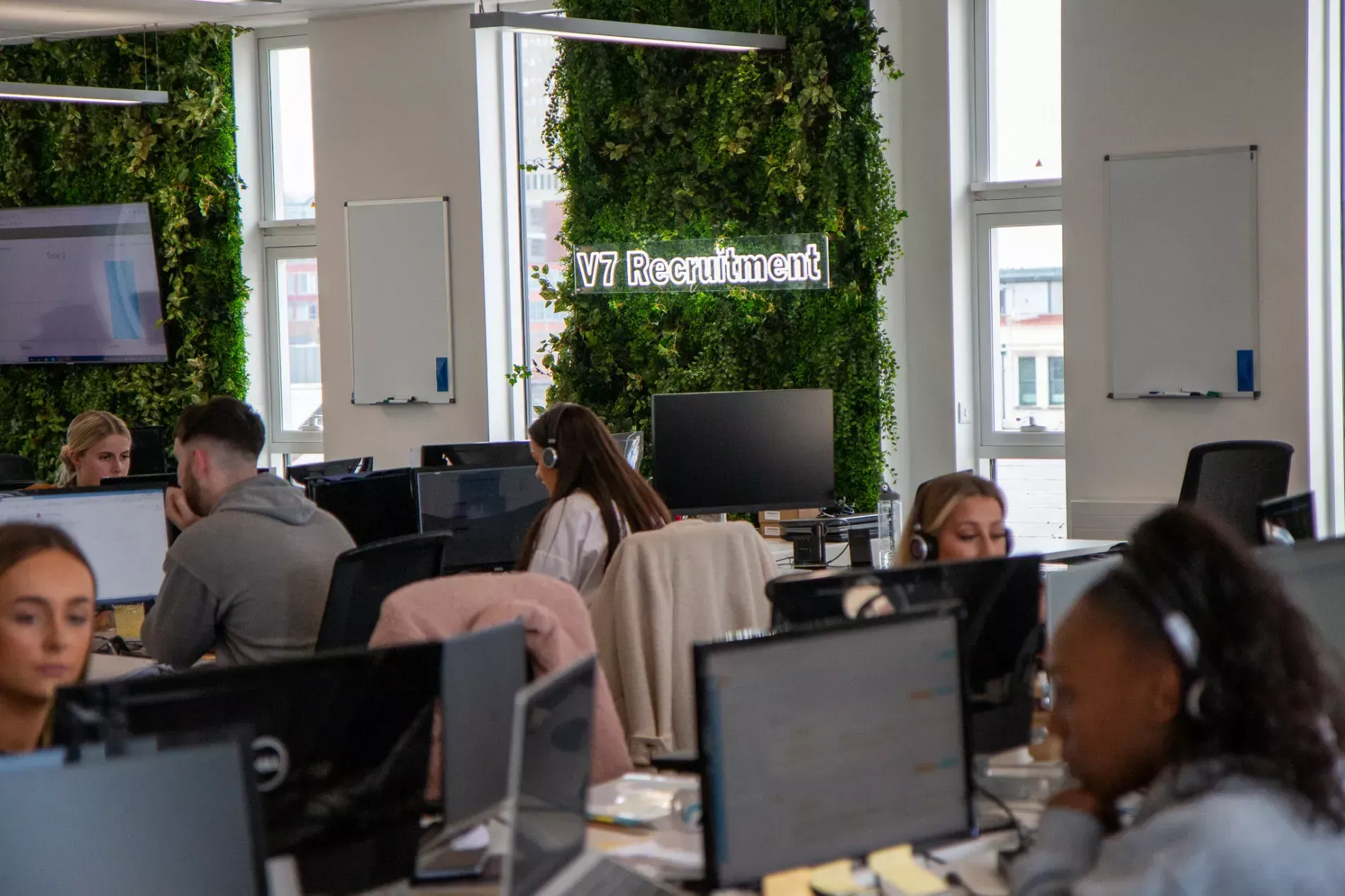 People working at computers in an office. Green plant wall in the background, logo visible.