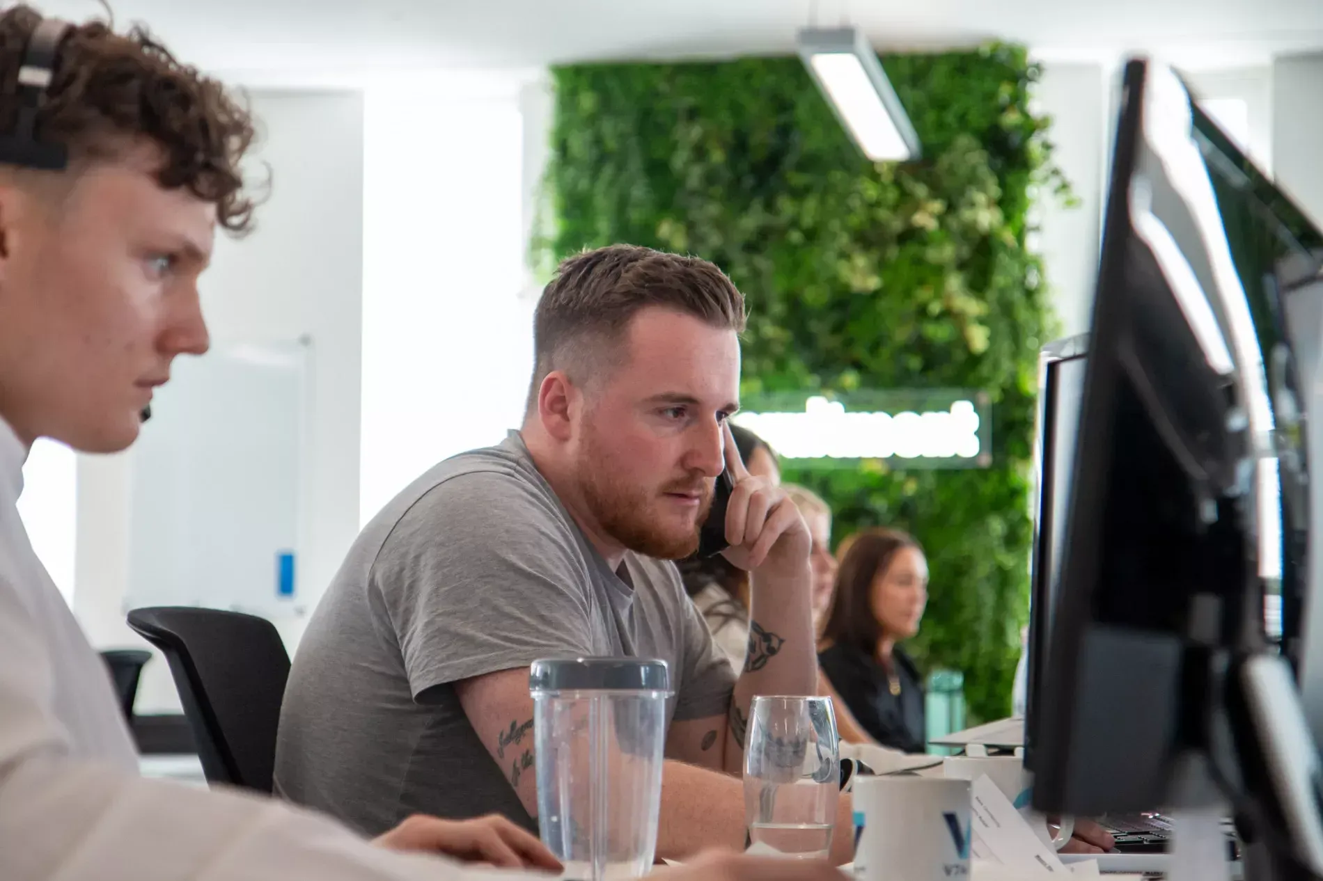 Two men working at computers in an office, one on the phone, with a wall of greenery.