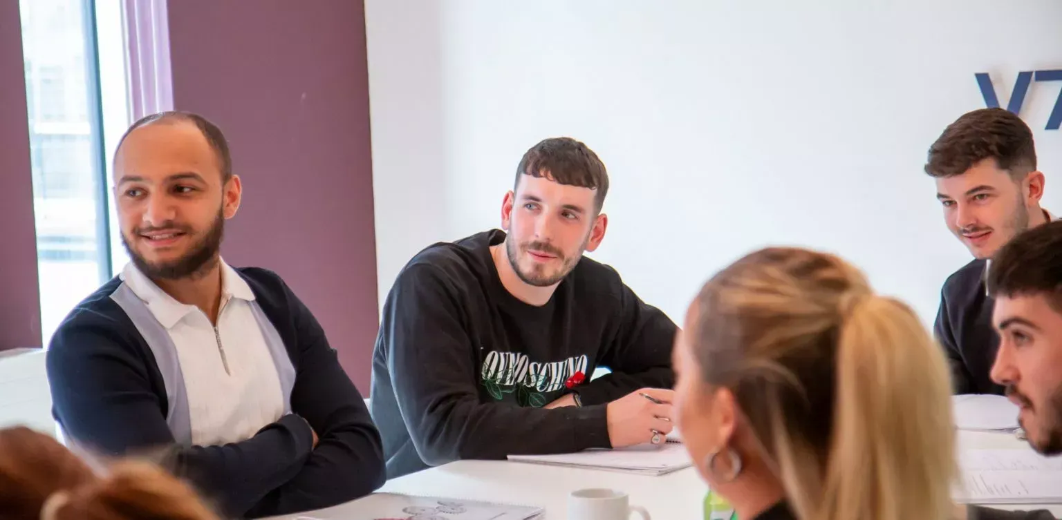 A group of people in a meeting room, discussing and looking at paperwork. Some have focused expressions.