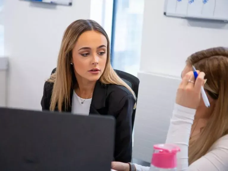 Two women in an office, one looking intently at a laptop, the other holding a pen, talking.