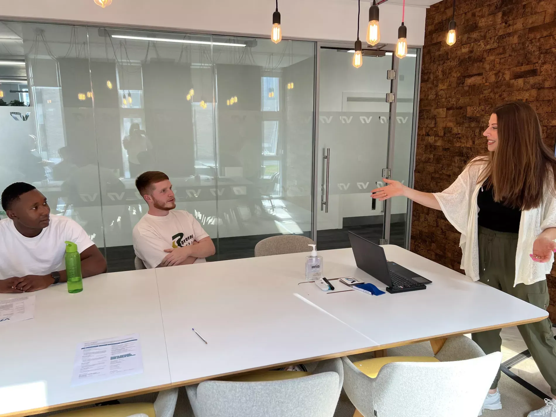 Three people in a modern office setting; a woman presents to two seated men at a table.