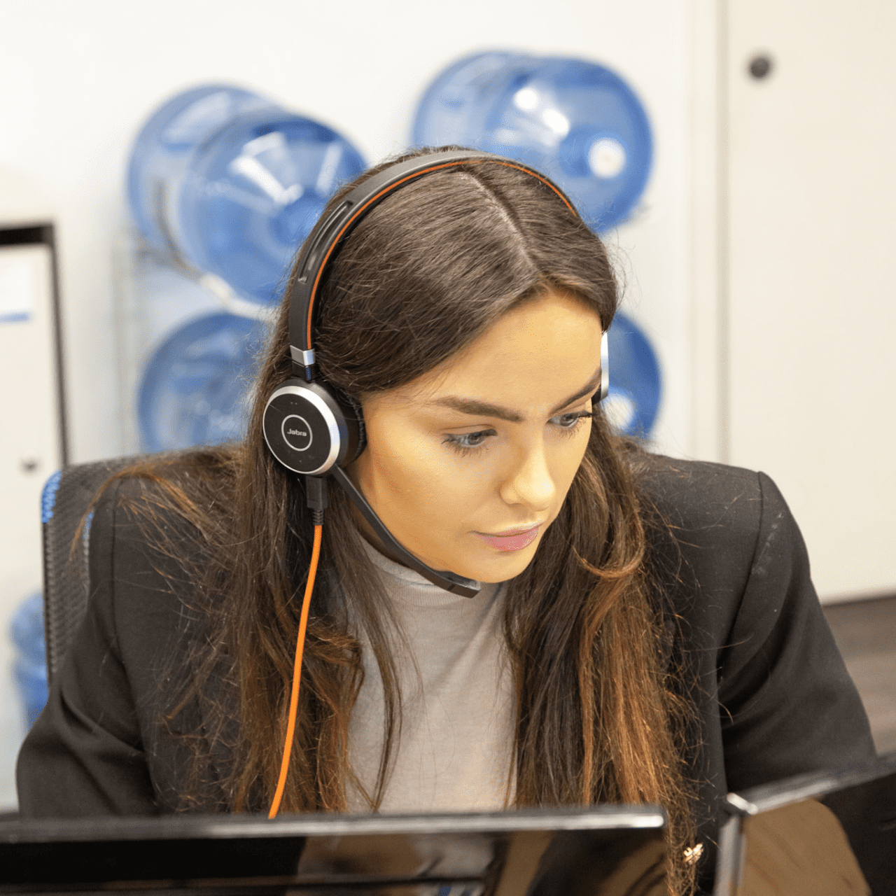 Woman wearing a headset, working at a desk. Behind her are several large water jugs.