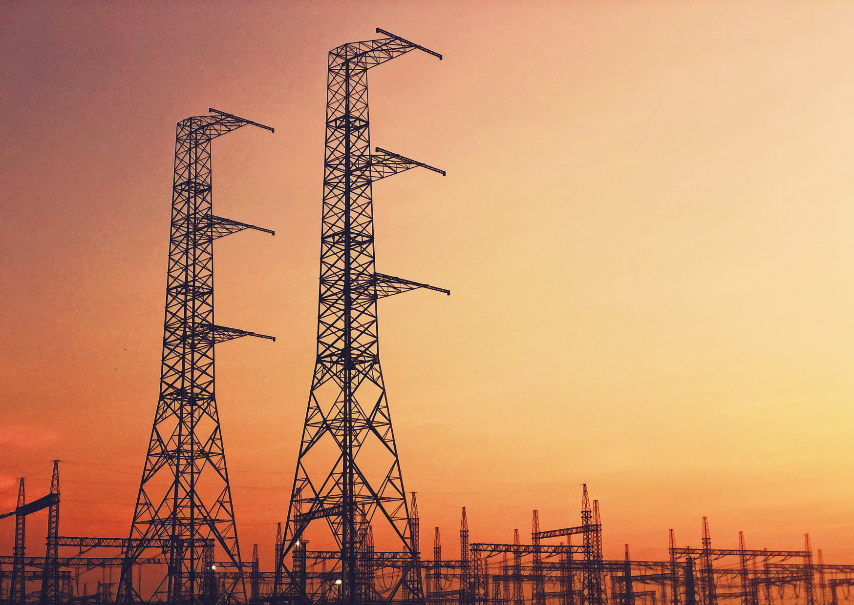 Two tall power line towers silhouetted against an orange and yellow sunset.