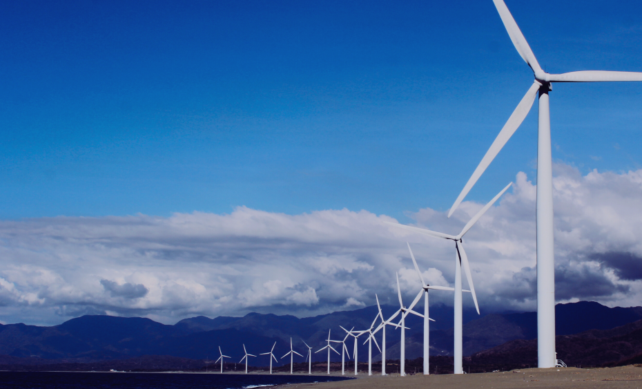 Wind turbines along a coast under a bright blue sky, with clouds and mountains in the background.