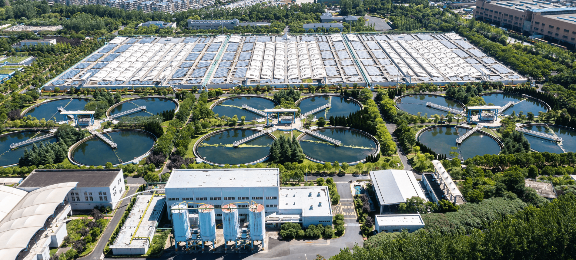 Aerial view of a water treatment plant with multiple circular ponds and large rectangular buildings surrounded by greenery.