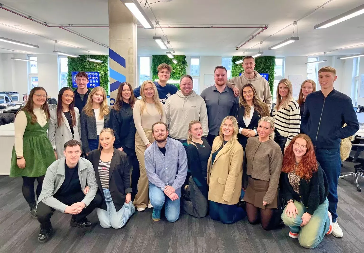 Group of young adults smiling in an office. Some are kneeling. Green plant wall in the background.